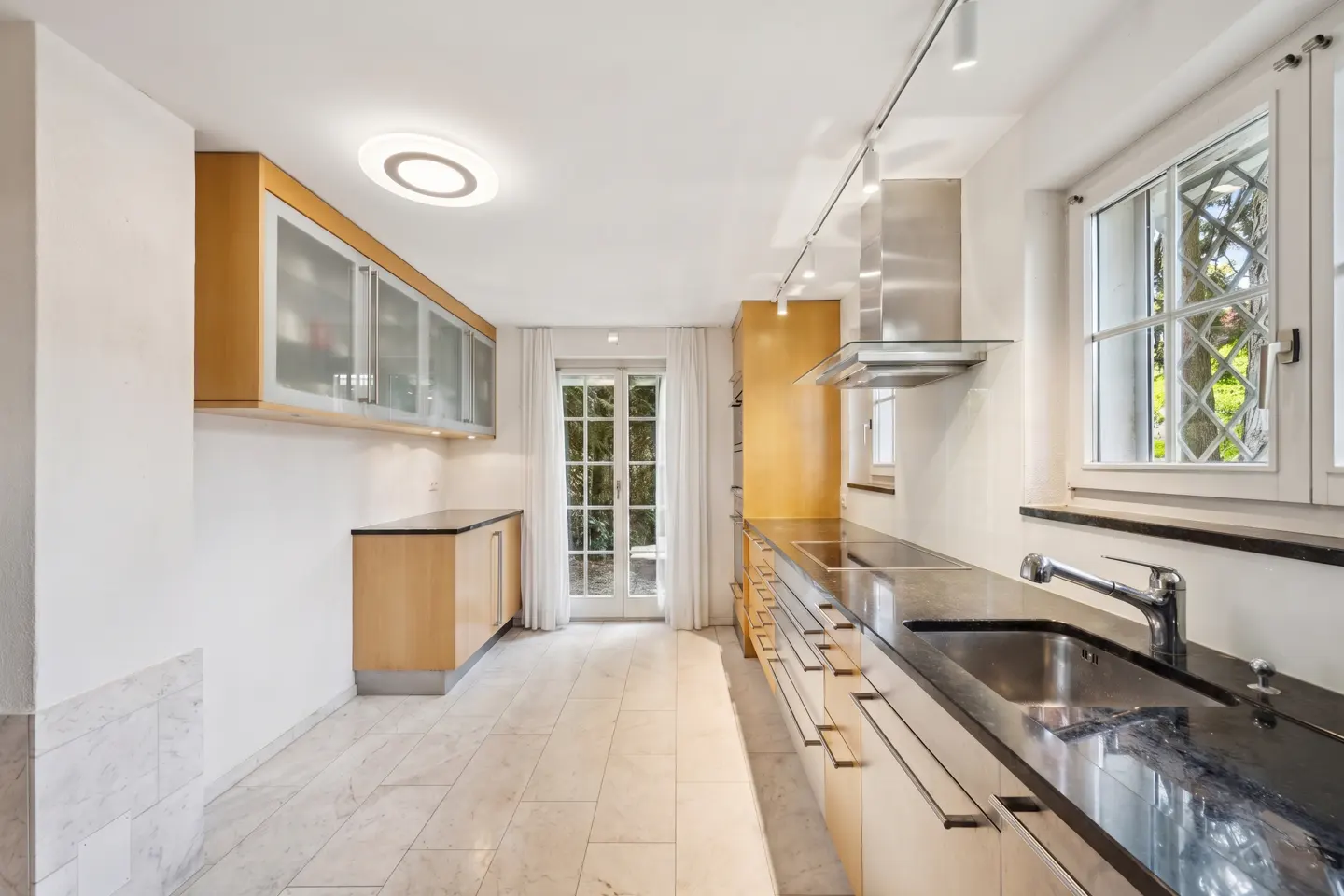 Bright kitchen with light wood cabinets, black countertops, and marble floors. A window and door let in natural light.