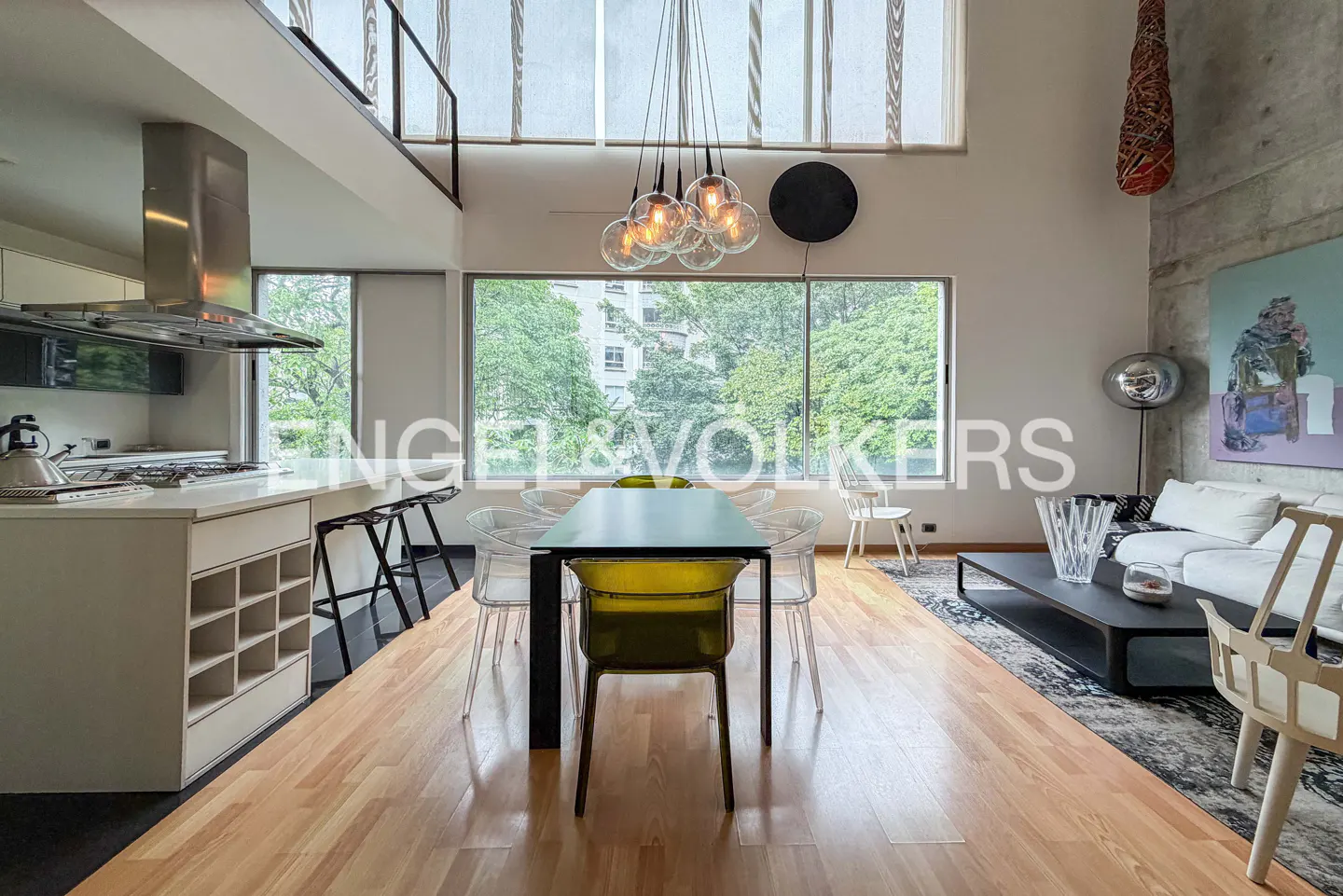 Open-concept living space with wood floors, a dining table with clear chairs, and a kitchen island. Large windows offer a view of greenery.