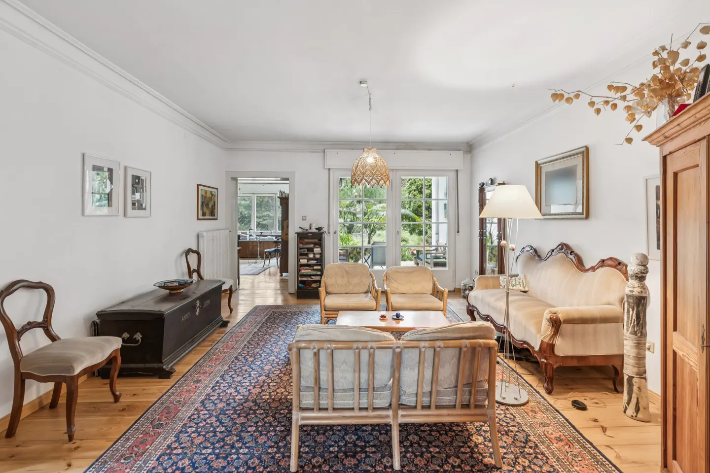 Bright living room with hardwood floors, a blue patterned rug, and antique furniture. Windows overlook a green garden.