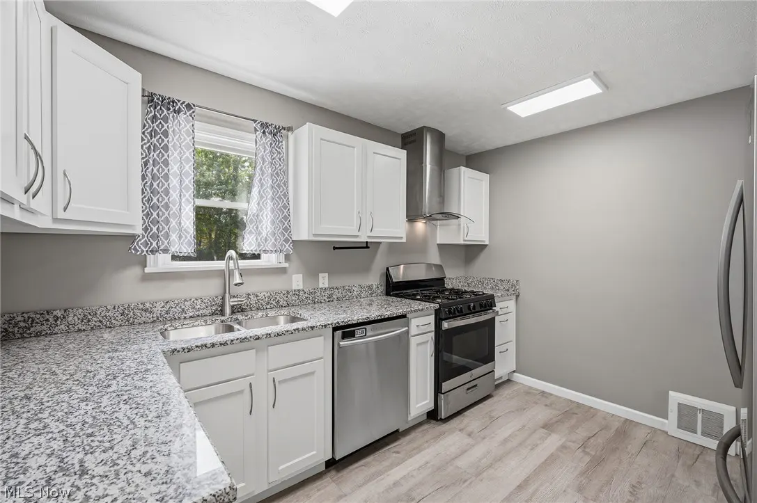 A bright kitchen with gray walls, white cabinets, stainless steel appliances, and granite countertops. A window with patterned curtains is above the sink.