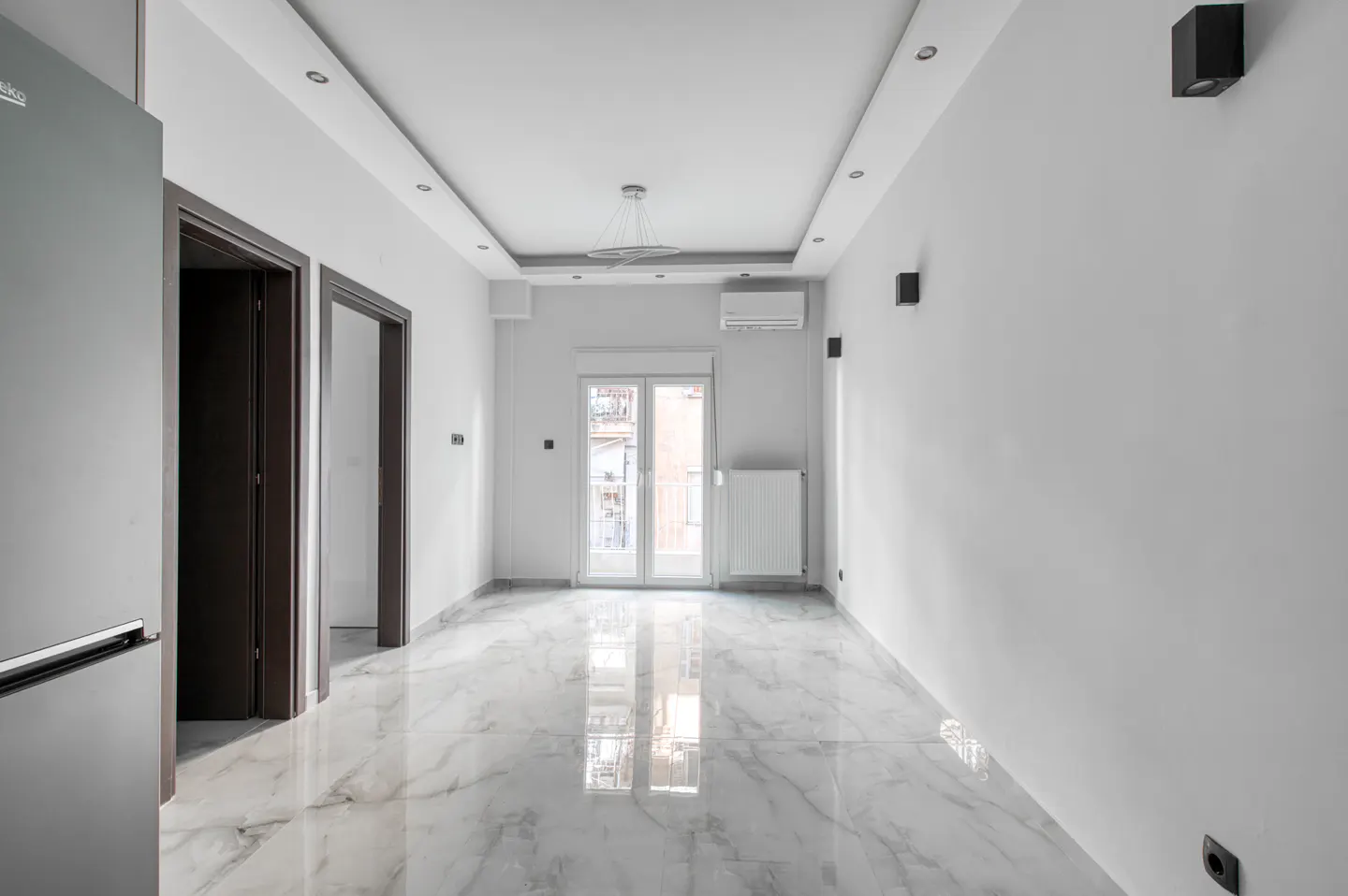 Bright, empty room with white walls, marble floors, and recessed lighting. A refrigerator and doorways are on the left. Double doors lead to a balcony.