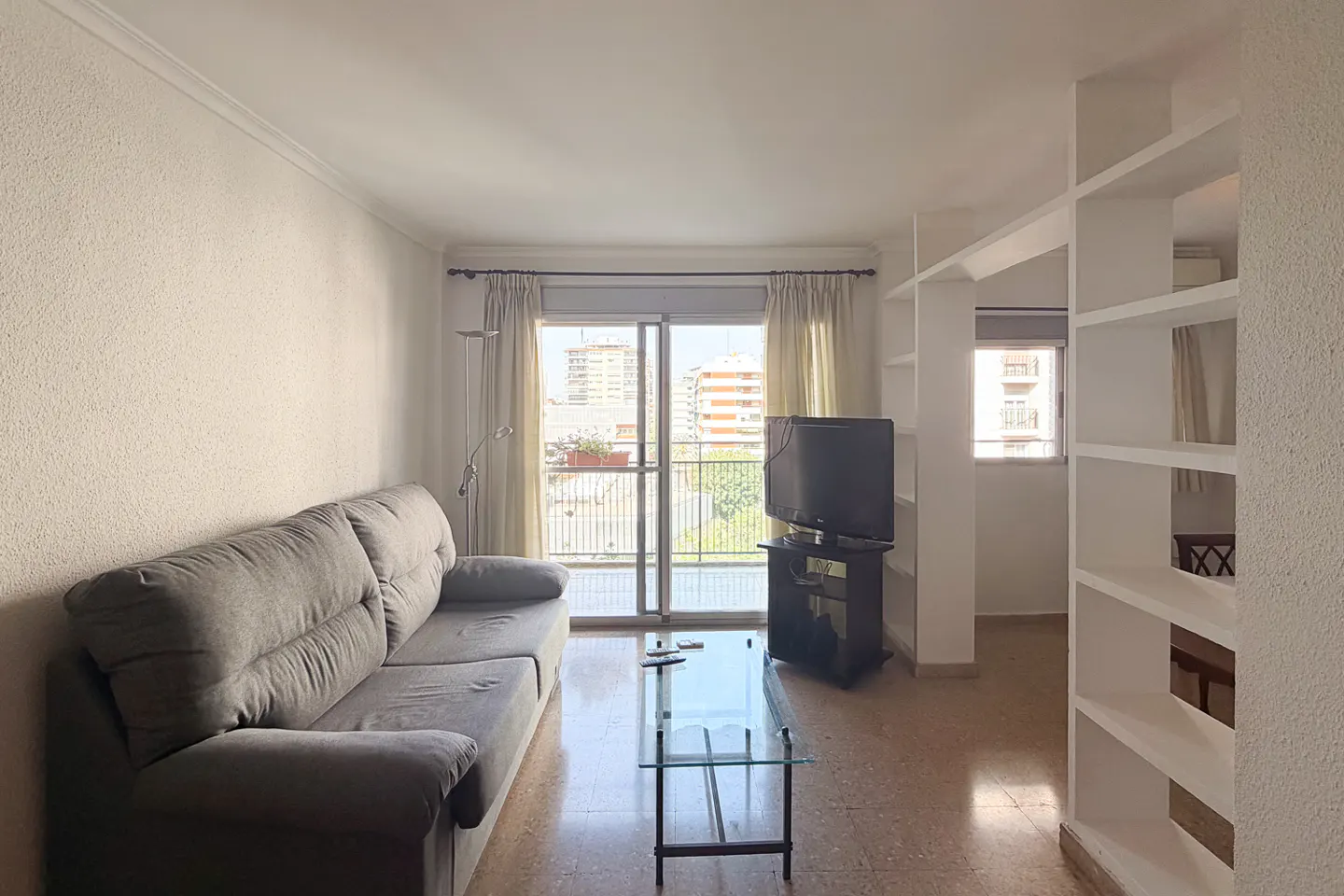Living room with a gray sofa, glass table, TV, and balcony with city view. White walls and built-in shelves.