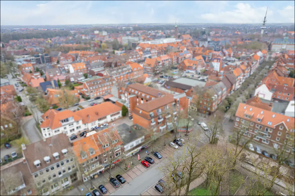 Aerial view of a European city with red-tiled roofs, streets with parked cars, and a distant tower under a cloudy sky.
