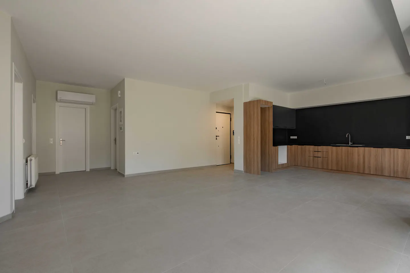 A bright, empty apartment with gray tile floors, white walls, and a modern kitchen with wood cabinets and a black backsplash.