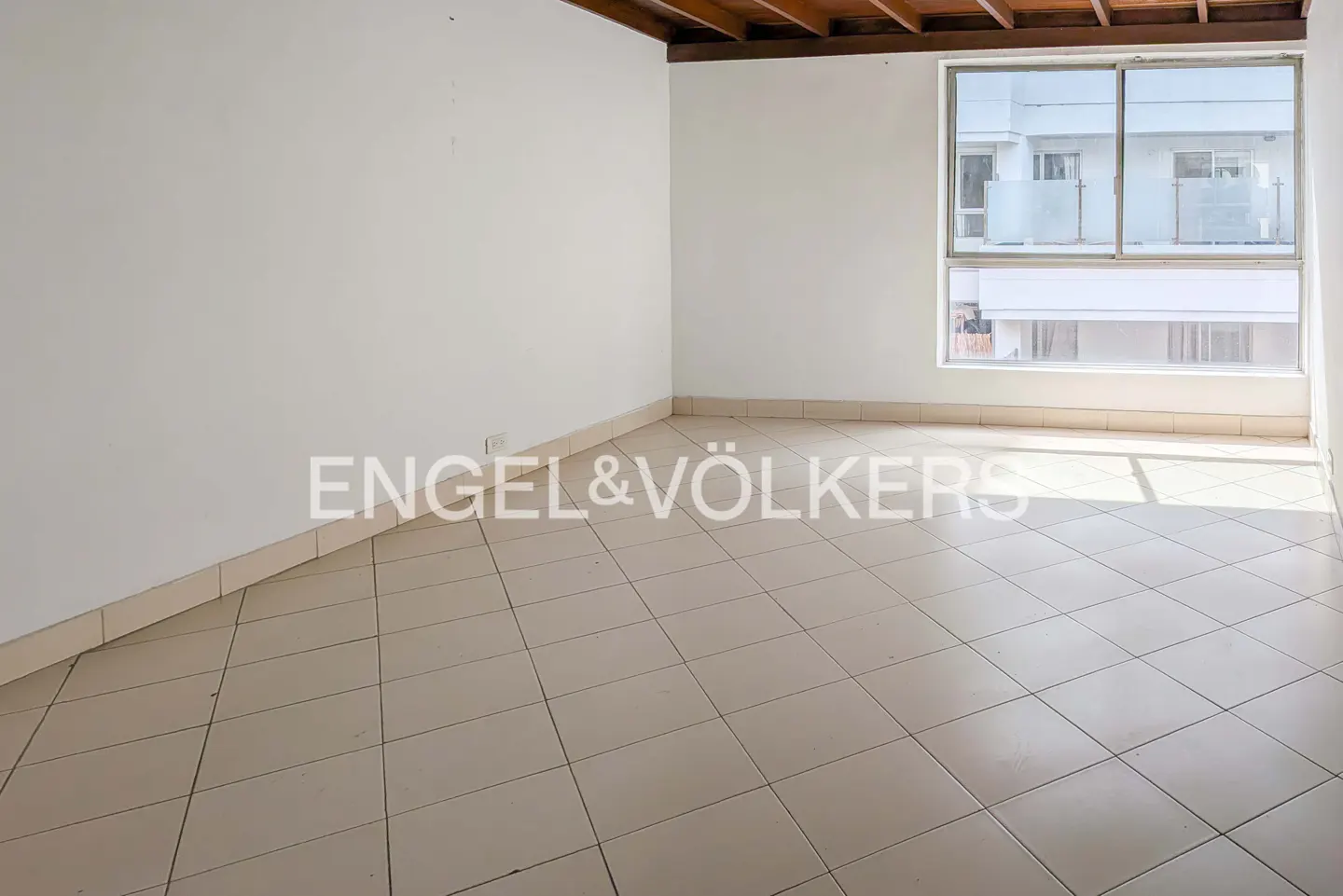 Empty room with white walls, tile floor, and wood ceiling. A large window shows a building outside. Engel & Völkers logo is visible.
