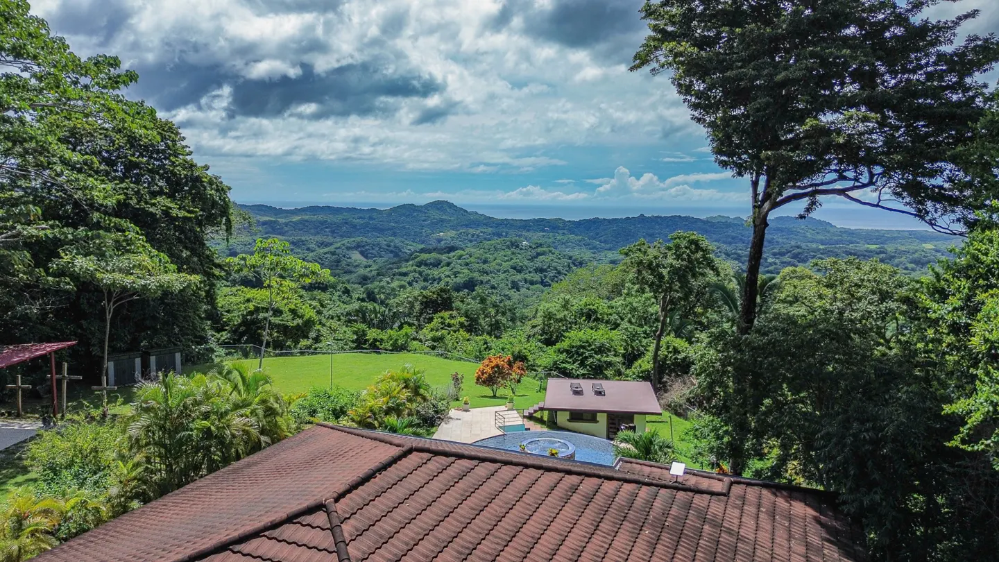 View from a rooftop with brown tiles, overlooking a lush green valley, a pool, and a small house. Cloudy sky in the background.