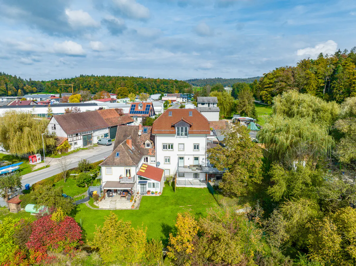 Aerial view of a white house with a red tile roof, surrounded by green lawn and trees in a rural setting.