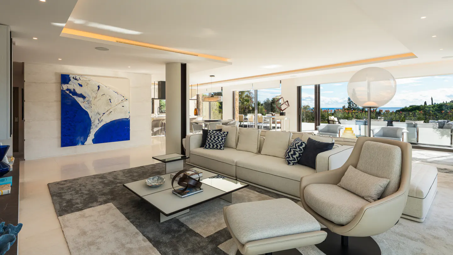 Bright, modern living room with a white sofa, chair, and a blue abstract painting. Large windows overlook a lush outdoor area.