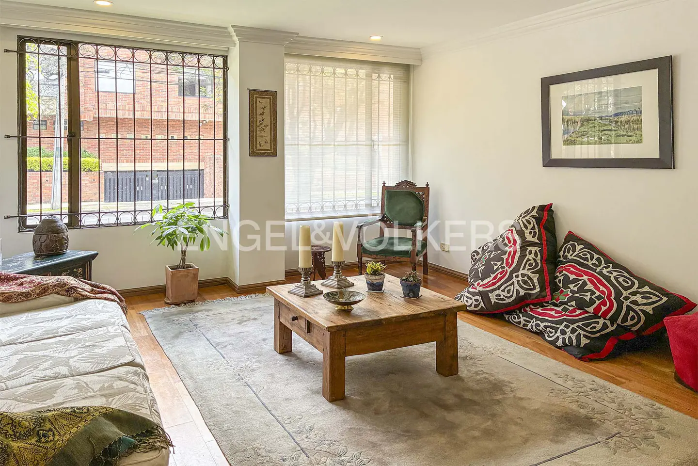 Living room with a wooden coffee table, patterned pillows, and a green chair. A large window with bars overlooks a brick building.