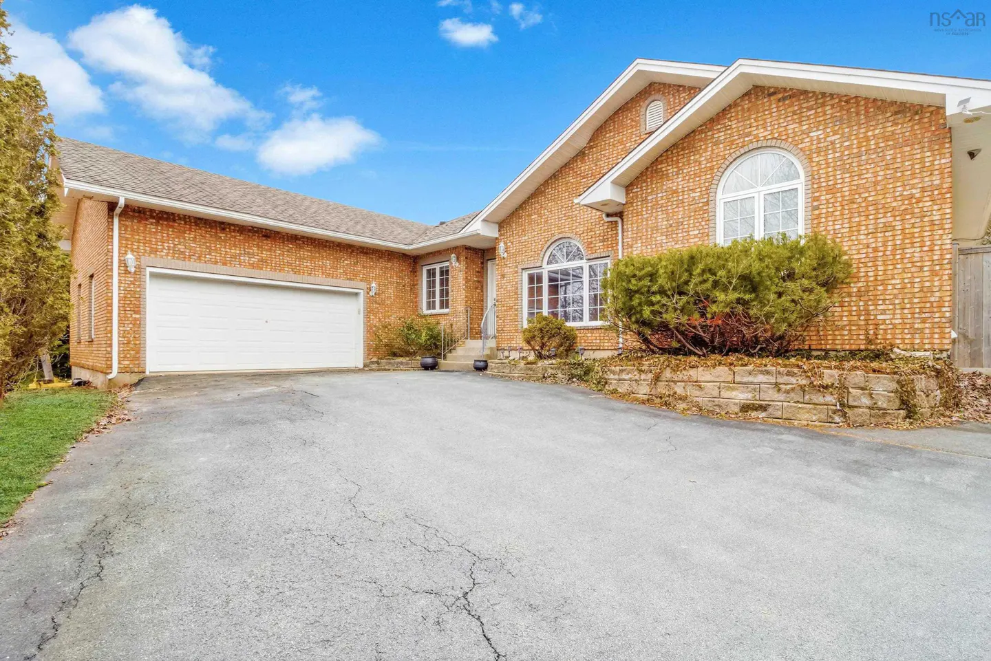 A one-story brick house with a white garage door and a gray driveway under a blue sky.