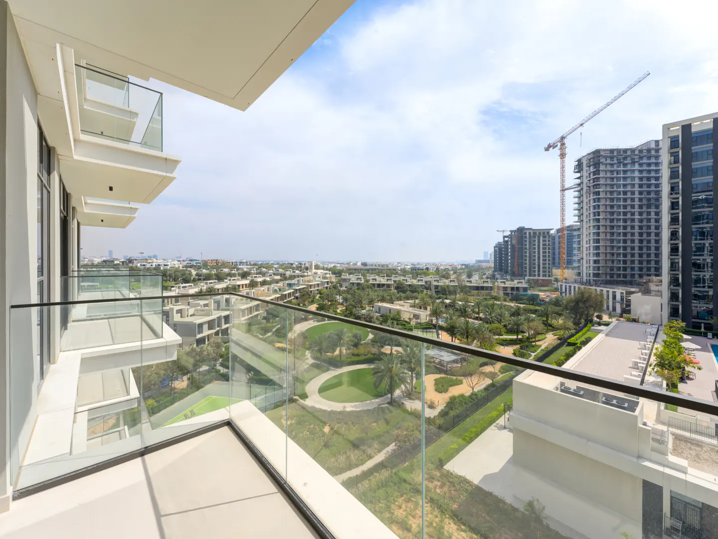 Balcony view of a green residential area with modern buildings and a construction crane under a blue sky. Glass railings are in the foreground.