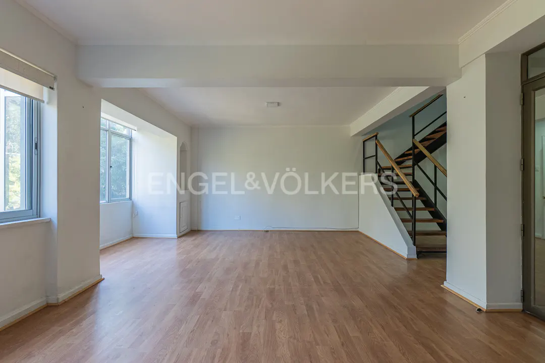 Bright, empty room with wood floors, white walls, and large windows. Stairs with black railings lead up. Engel & Völkers logo on the wall.