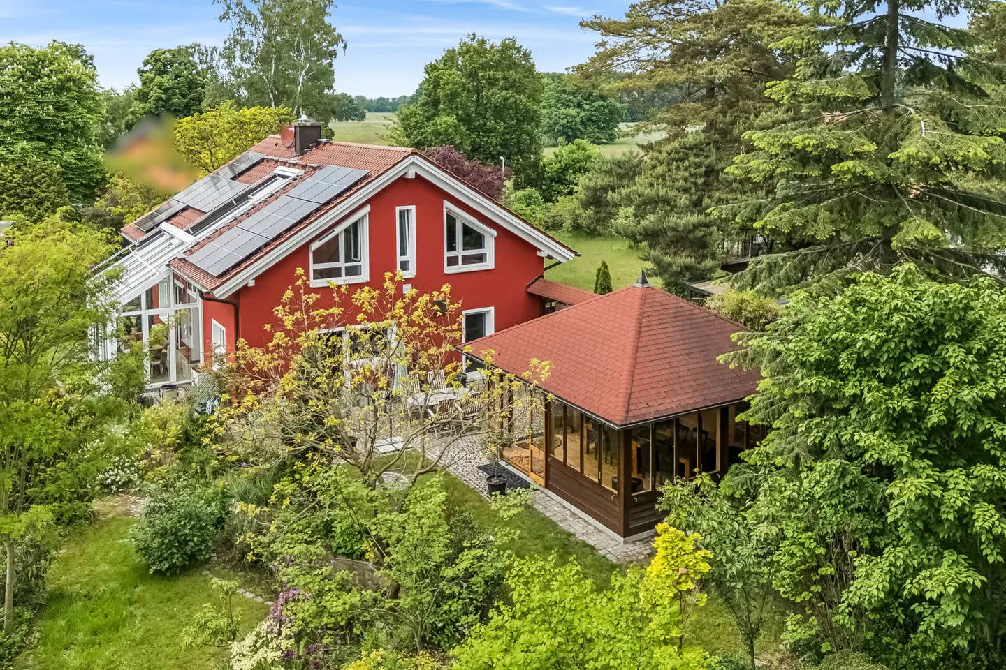A bright red house with solar panels and a glass sunroom, featuring a separate wooden gazebo in a lush, green garden.