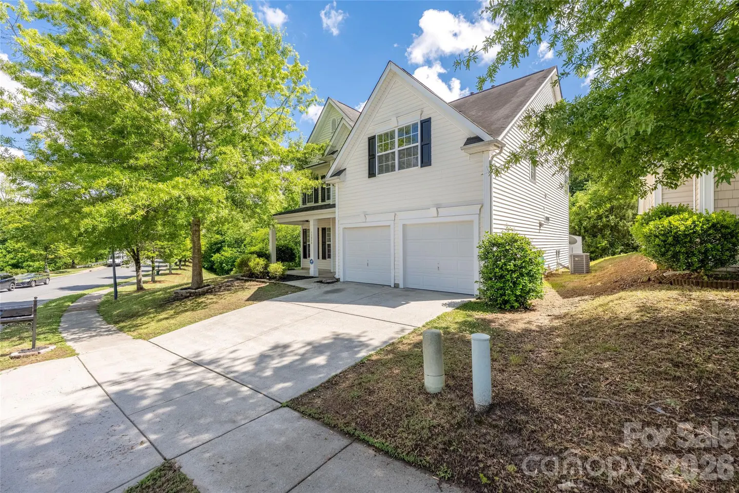 Two-story white vinyl siding house with a two-car garage, concrete driveway, and lush green trees under a blue sky.