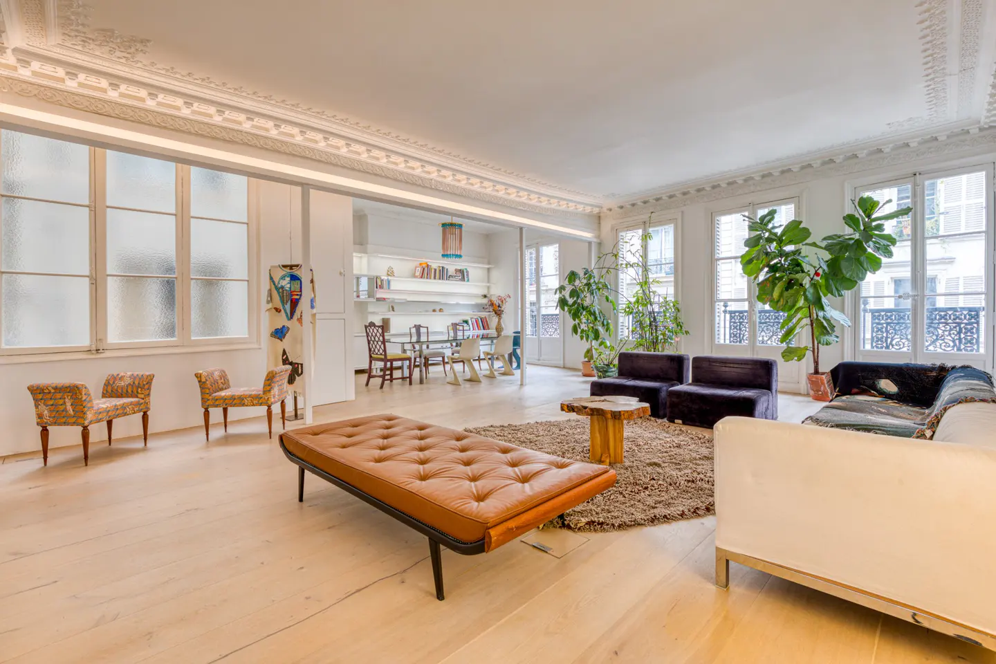 Bright living room with hardwood floors, large windows, and ornate ceiling trim. Furnished with a brown leather bench, sofas, and indoor plants.