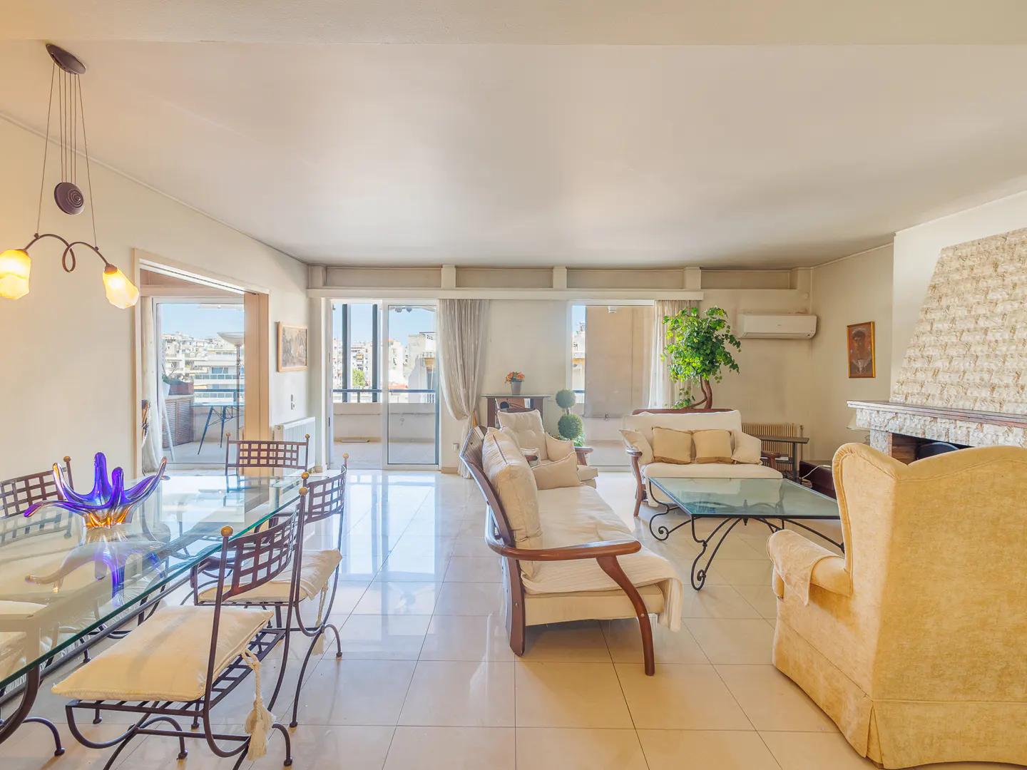 Bright, open-plan living and dining area with glass table, sofas, and balcony access. Cream walls and tile floors.