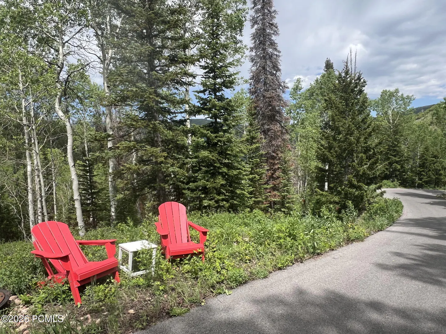 Two red Adirondack chairs and a white table sit near a road, surrounded by lush green trees.