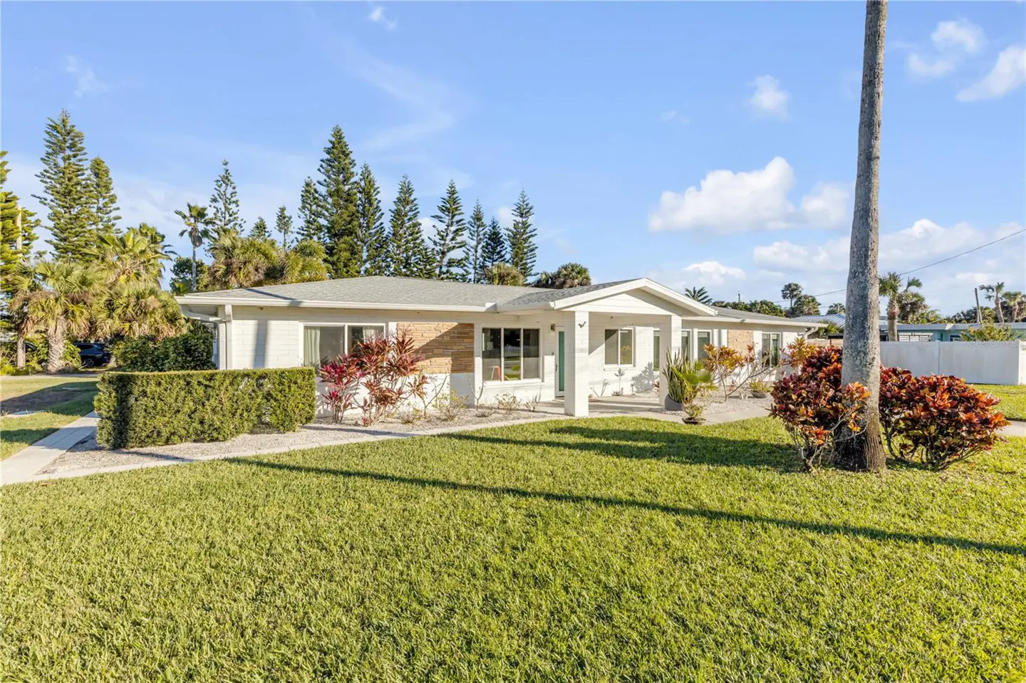 A single-story white house with a green lawn, bushes, and trees under a blue sky.