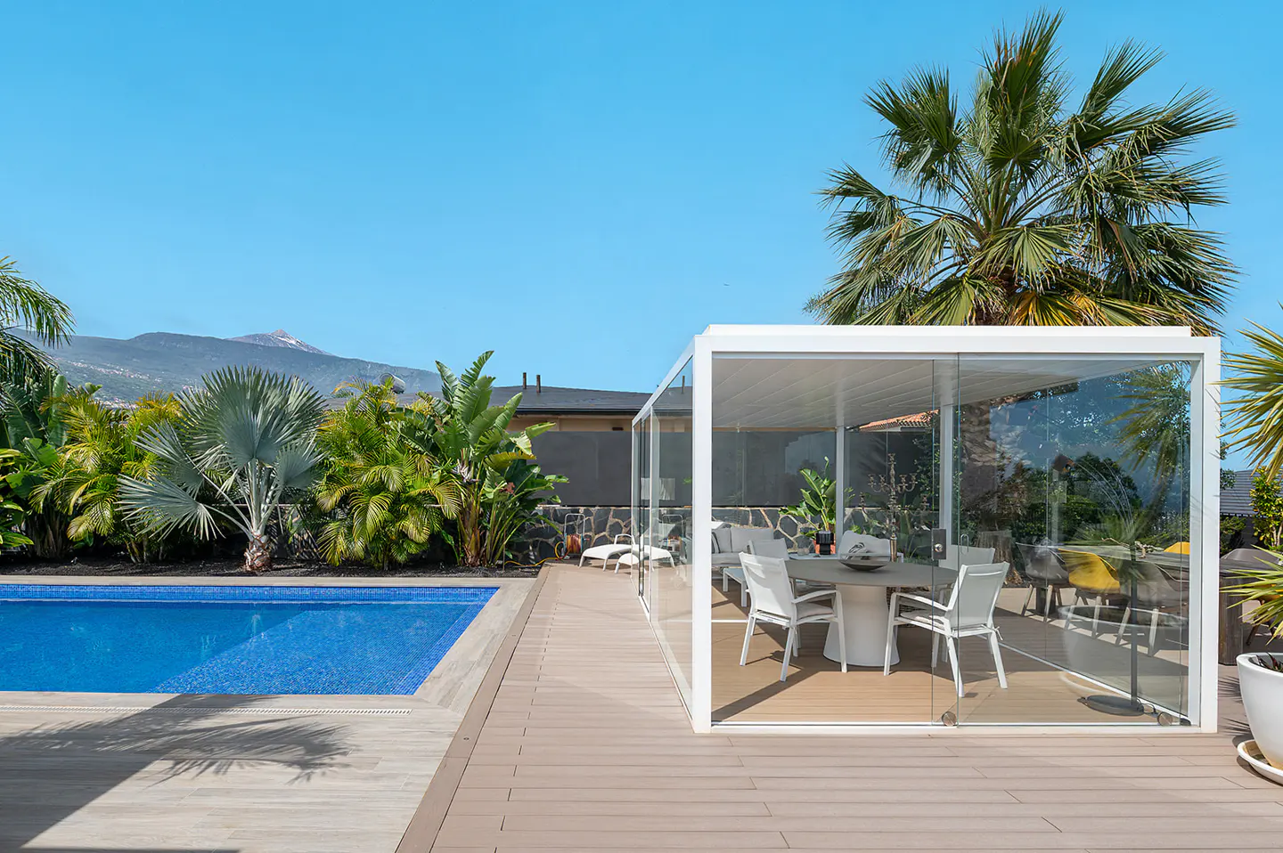 Outdoor patio with a pool, palm trees, and a glass-enclosed dining area with white chairs and a table. Blue sky.