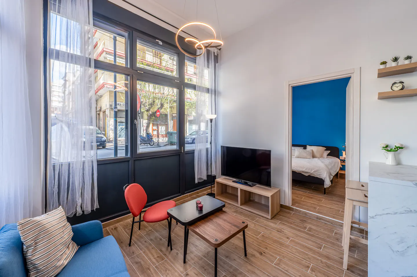 Bright apartment interior with a blue sofa, red chair, and TV. A bedroom with blue walls is visible through a doorway. Large windows show a city street.