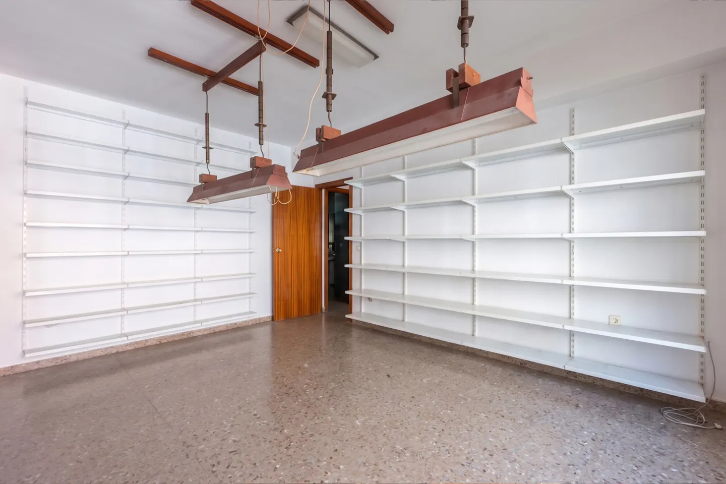 Empty room with white walls, shelves, and terrazzo floor. Two brown industrial lights hang from the ceiling. A wooden door is visible in the background.