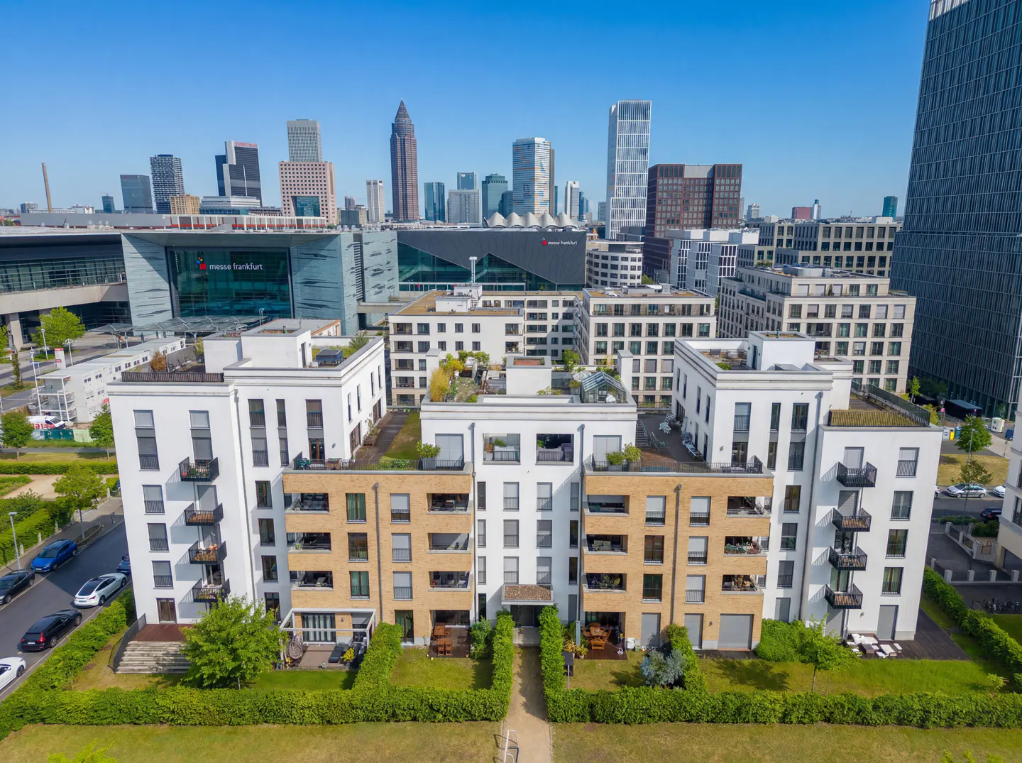 Aerial view of modern apartments with white and tan facades, green lawns, and a city skyline in the background.