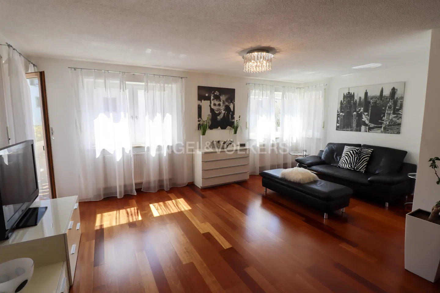 Bright living room with hardwood floors, white walls, and sheer curtains. A black leather sofa and ottoman sit near a white dresser with artwork above.