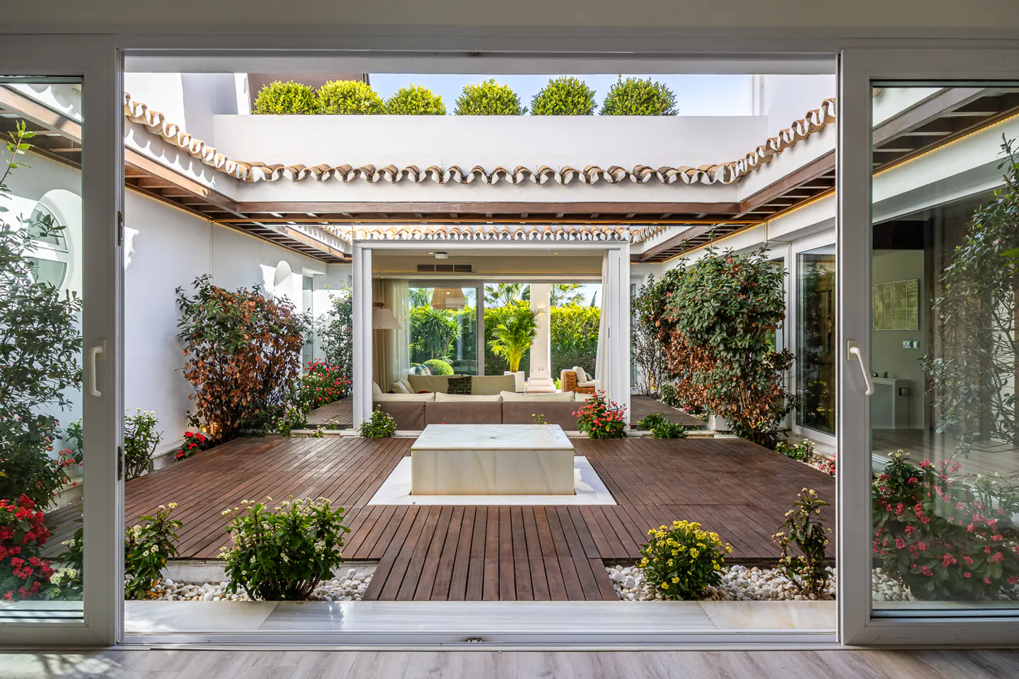View through open sliding doors to a courtyard with wood deck, white walls, and lush greenery. A white marble fountain sits in the center.