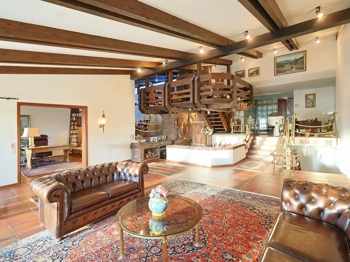 Living room with brown leather sofas, glass table, and red patterned rug. Wooden beams on ceiling and a unique wooden structure above a brick fireplace.