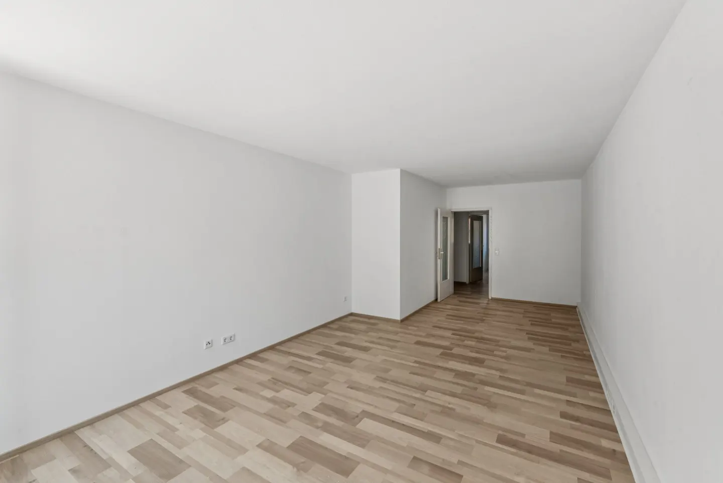 Empty room with white walls, light wood parquet flooring, and an open door leading to another room.
