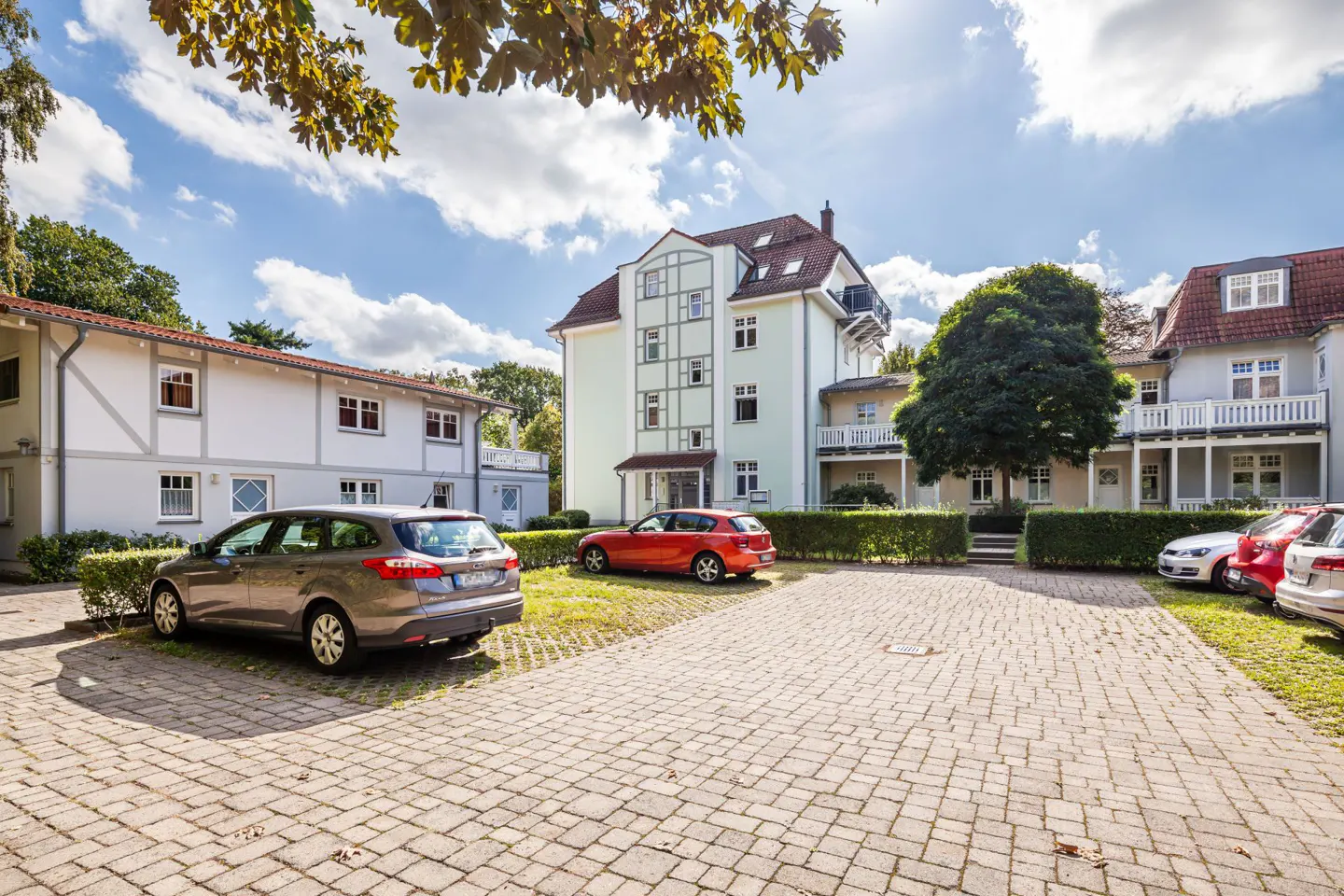 Exterior view of a light-colored apartment building with a brick parking lot and several parked cars.