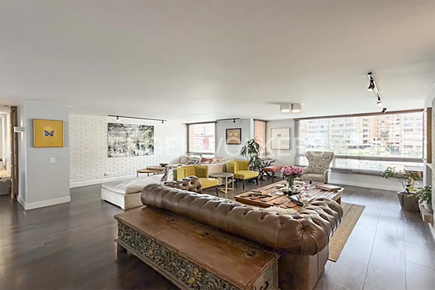 Wide shot of a living room with dark wood floors, a brown leather sofa, yellow chairs, and large windows.