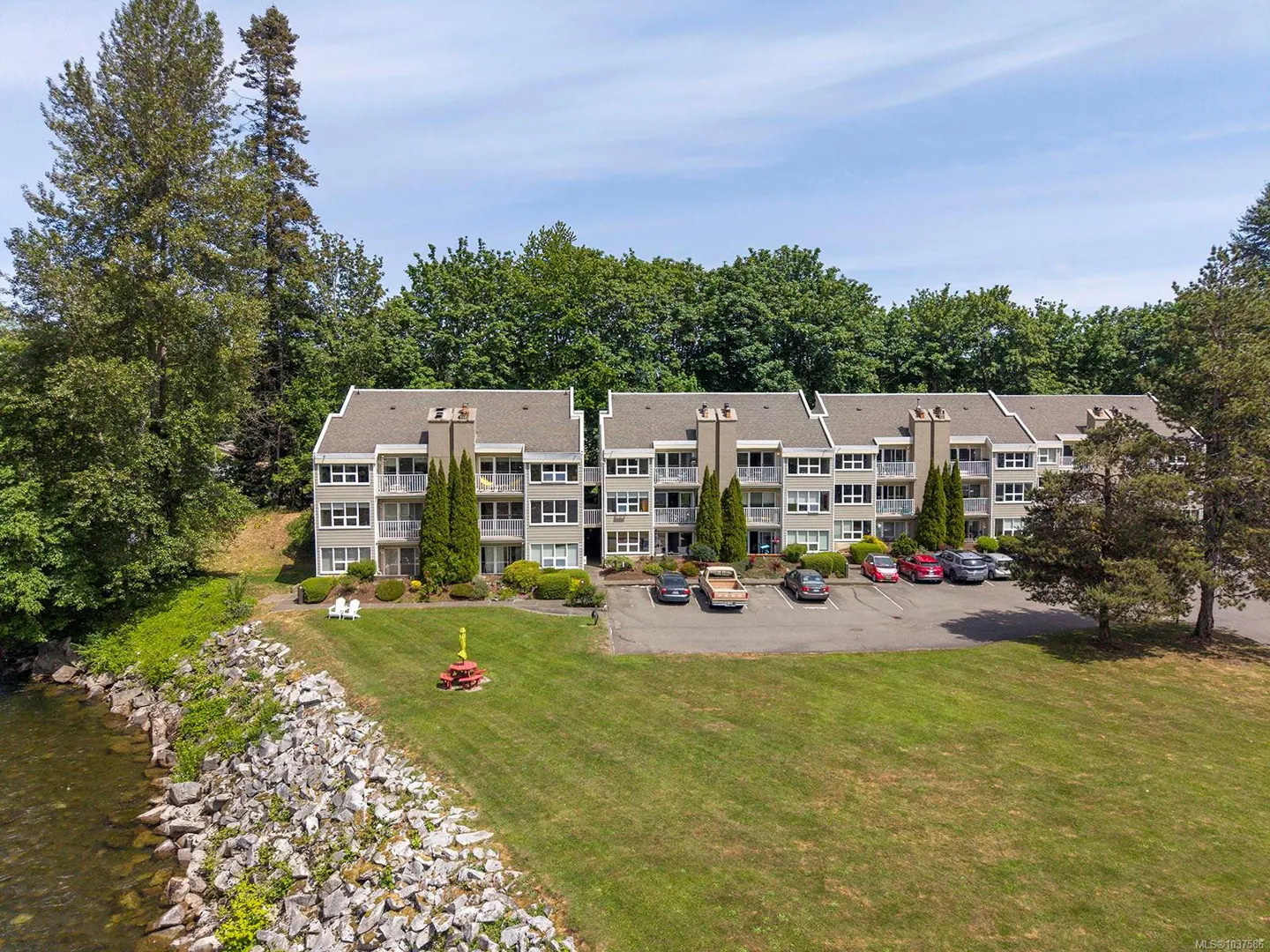 Aerial view of a three-story apartment complex with a parking lot, lawn, and waterfront. Cars are parked in front of the building. Trees surround the property.