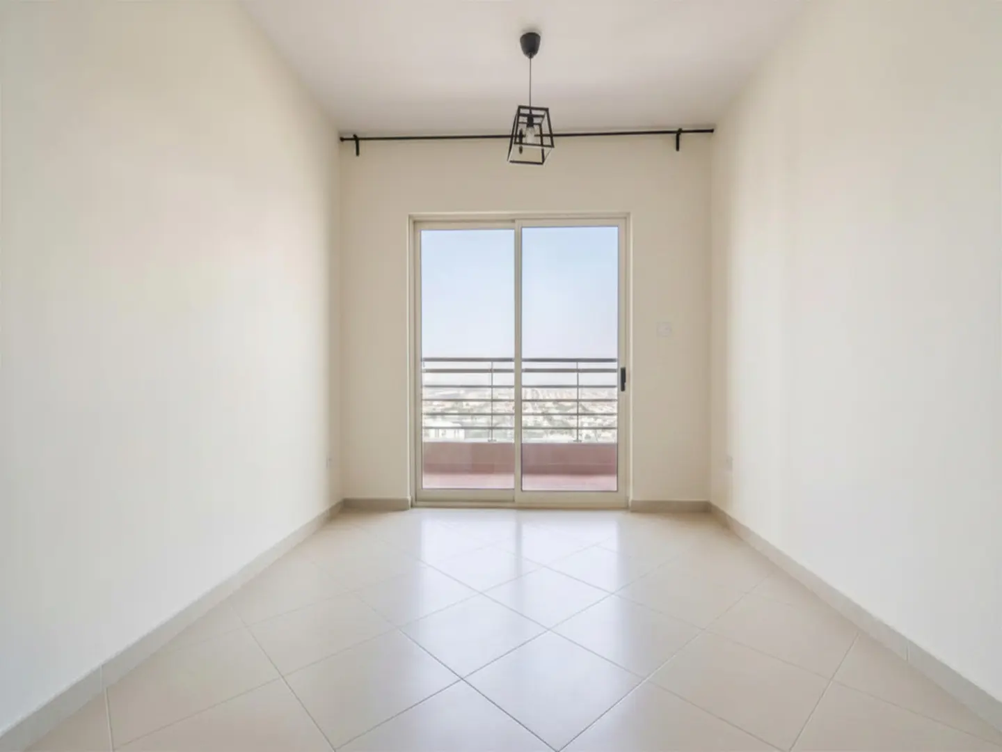 An empty room with white walls, tiled floor, and sliding glass doors to a balcony with a city view. A modern light fixture hangs from the ceiling.