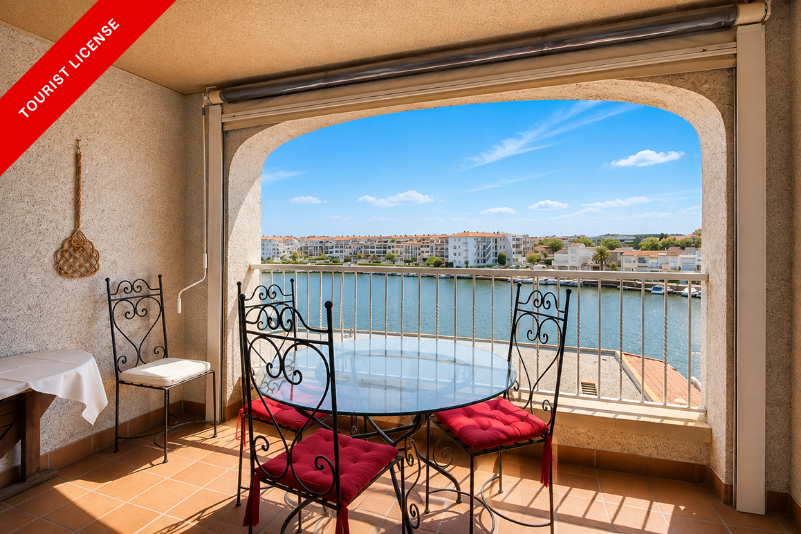 Balcony with a glass table and black metal chairs with red cushions, overlooking a canal and coastal buildings.