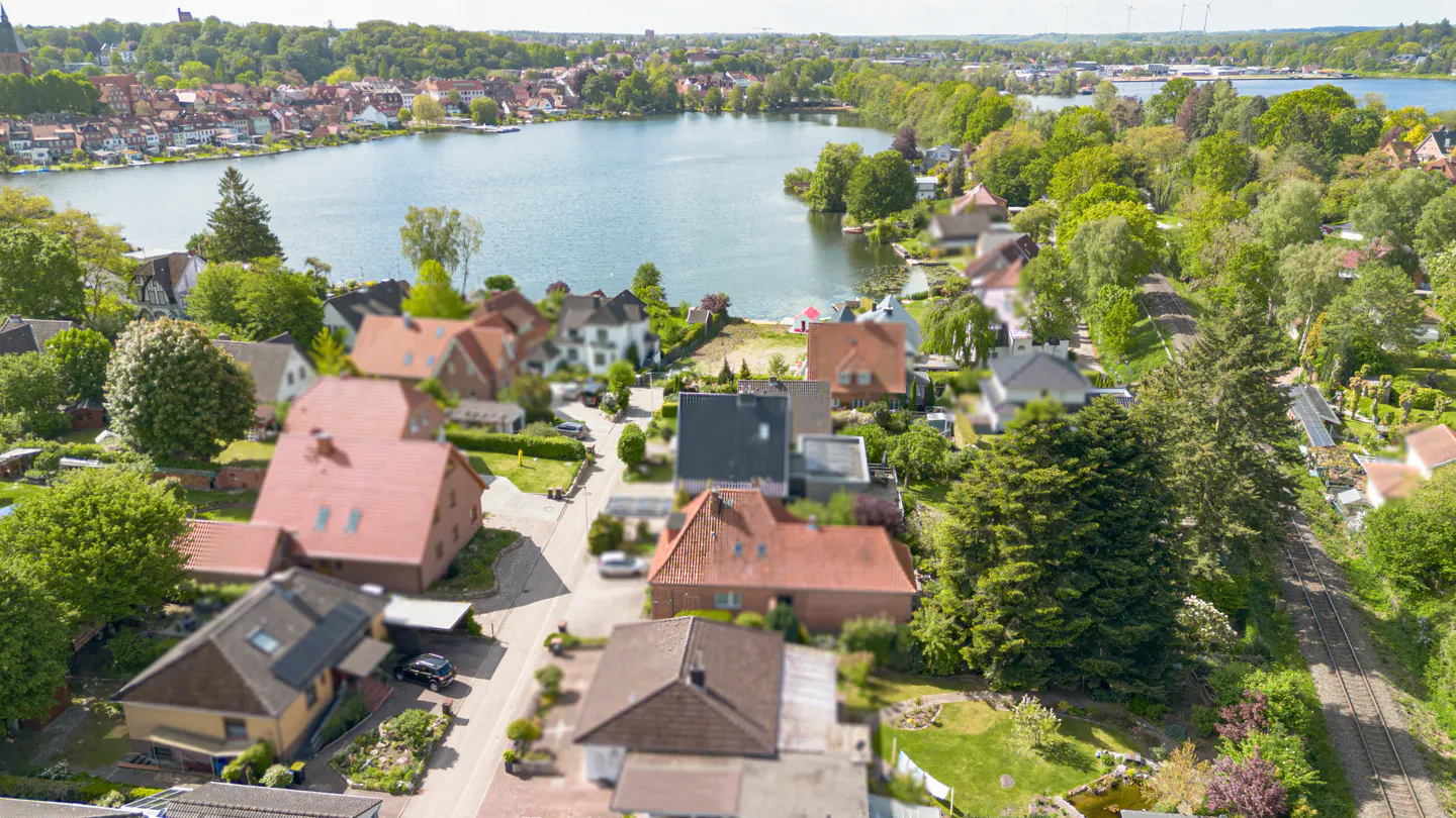 Aerial view of a residential neighborhood with red-roofed houses, lush green trees, and a scenic lake in the background.