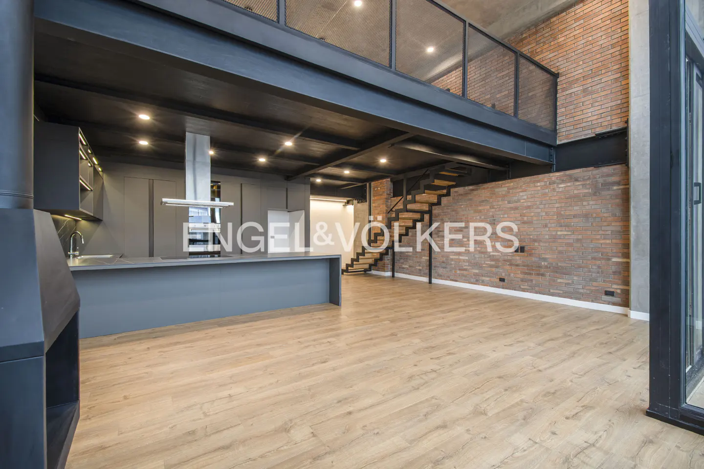 Modern industrial loft with a gray kitchen, wood floors, exposed brick walls, and a metal staircase to a mezzanine level.