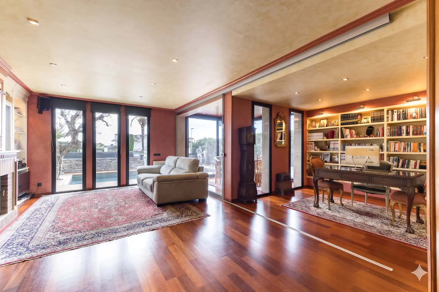 Open-concept living room with hardwood floors, a beige sofa, and a desk with bookshelves. Sliding glass doors lead to a pool.