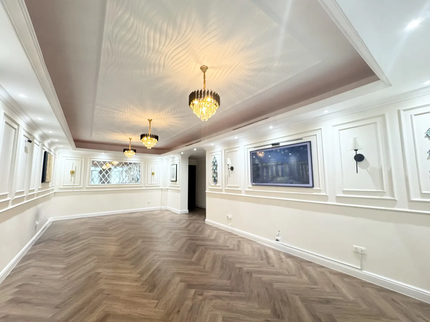 Spacious room with herringbone wood floors, white walls with decorative molding, and crystal chandeliers.