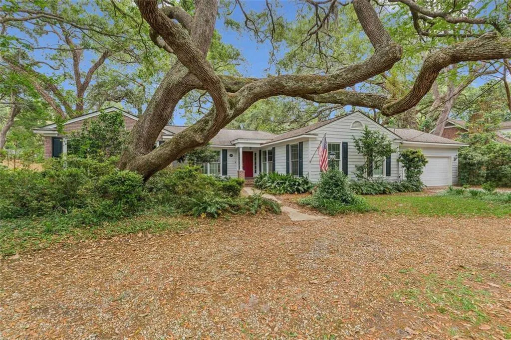 A white house with a red door and an American flag is shaded by a large tree.
