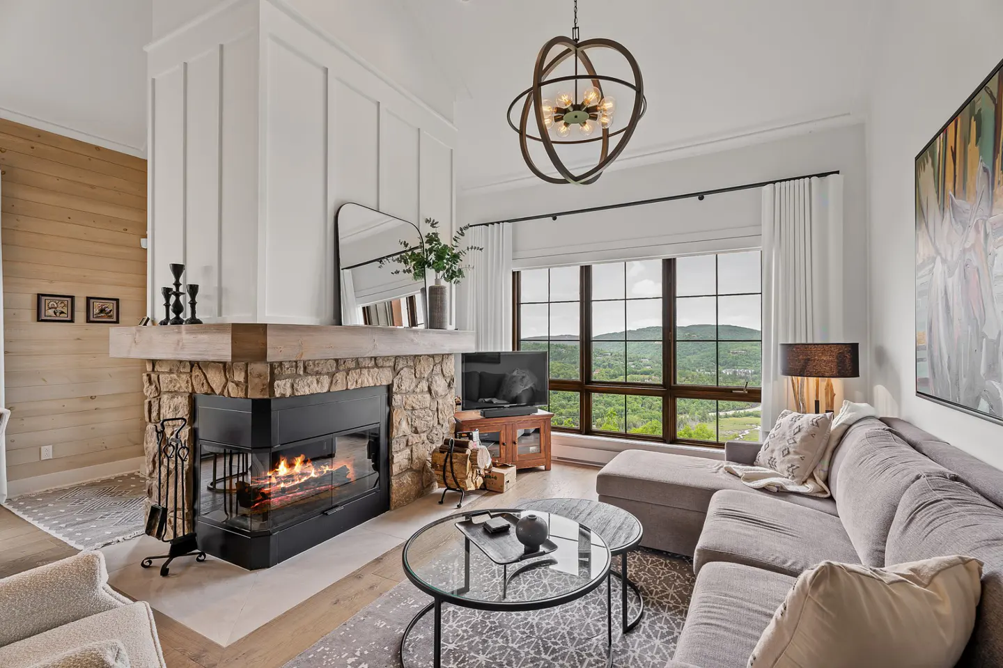 Living room with stone fireplace, gray sectional sofa, and large window with mountain views. A modern chandelier hangs from the ceiling.