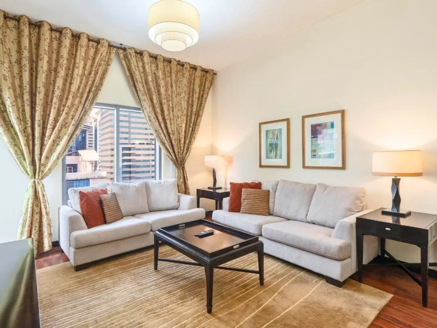 Living room with two sofas, a dark wood coffee table, and patterned curtains framing a city view. Two framed pictures hang on the wall.