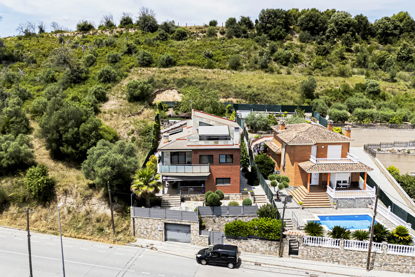 Aerial view of two brick houses nestled against a lush, green hillside, with a swimming pool and a black car parked below.