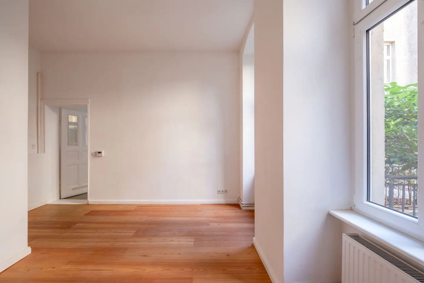Bright, empty room with hardwood floors and white walls. A window shows greenery outside. A doorway leads to another room.