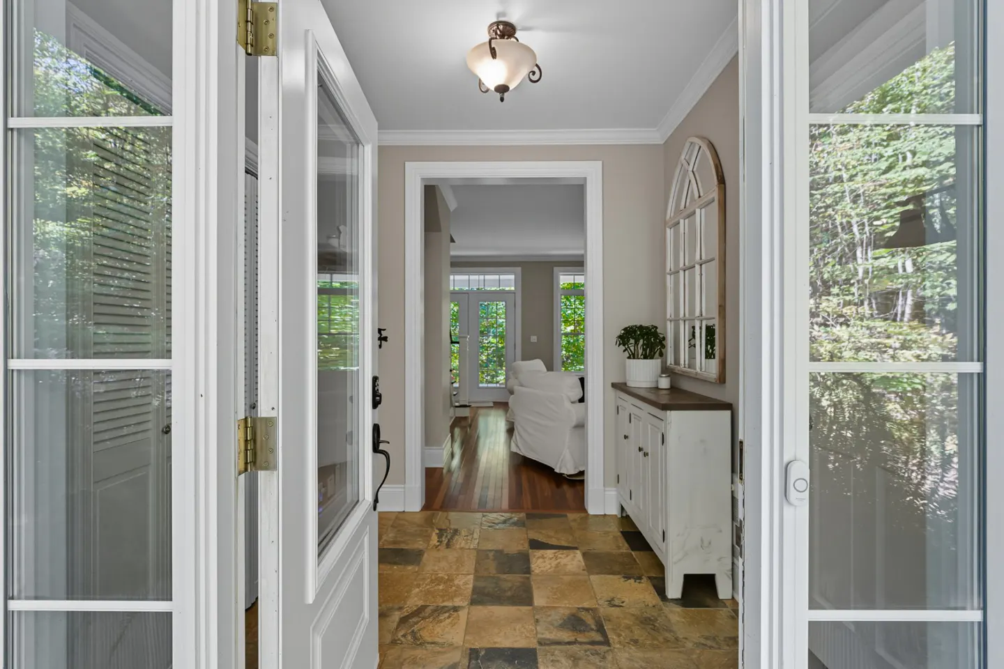 Open white french doors reveal a foyer with a stone tile floor, white cabinet, and arched mirror. A glimpse of a living room with a white sofa is visible.