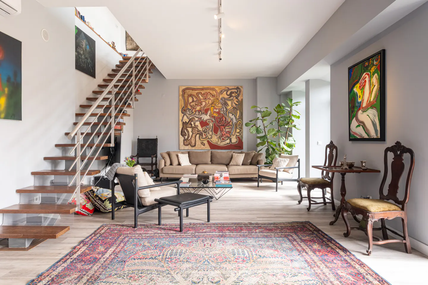A modern living room with a brown sofa, chairs, a colorful rug, and artwork on gray walls. A wooden staircase is on the left.