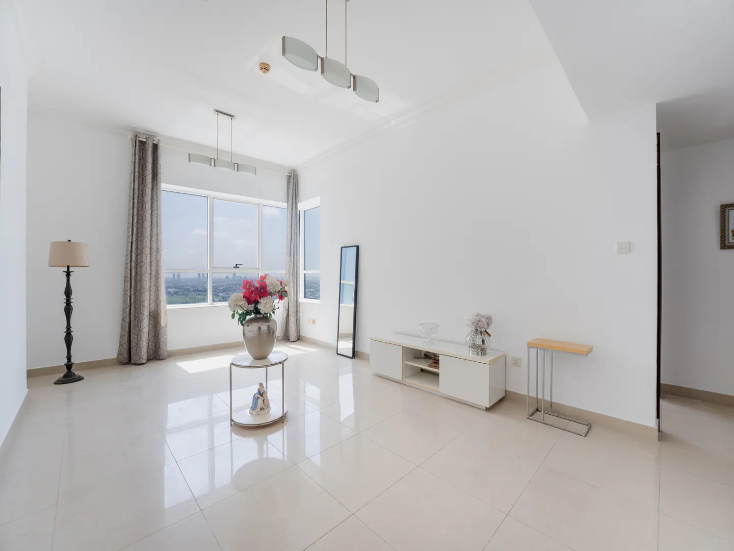 Bright, modern living room with white walls, tile floor, and large windows with city views. Features include a lamp, flowers, and minimalist furniture.
