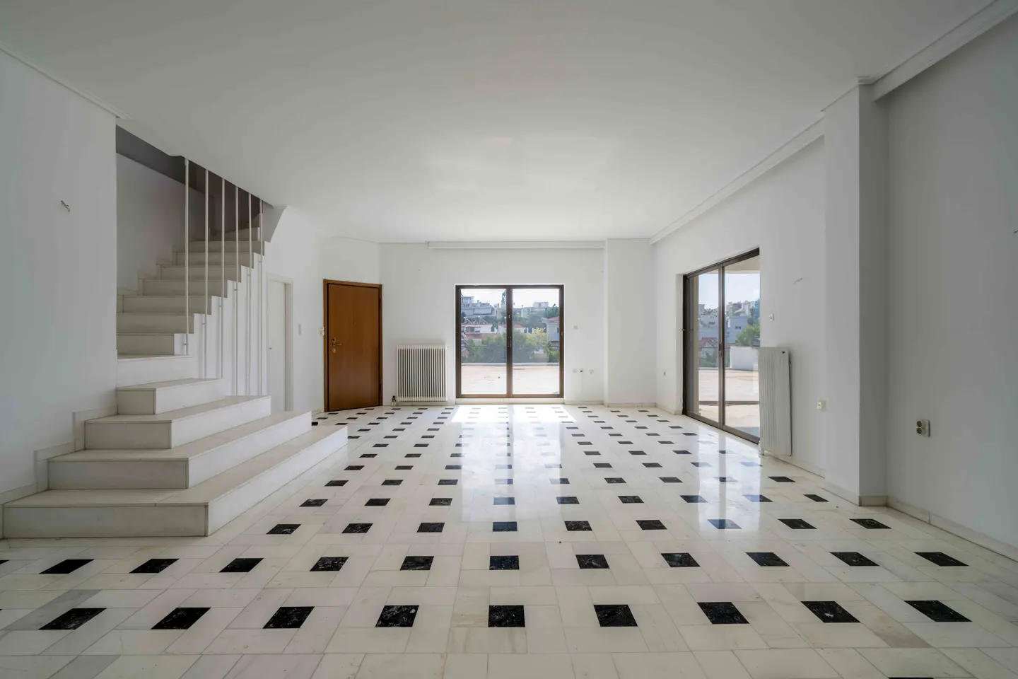 Bright, empty room with white walls, black and white tiled floor, stairs, and windows overlooking a cityscape.