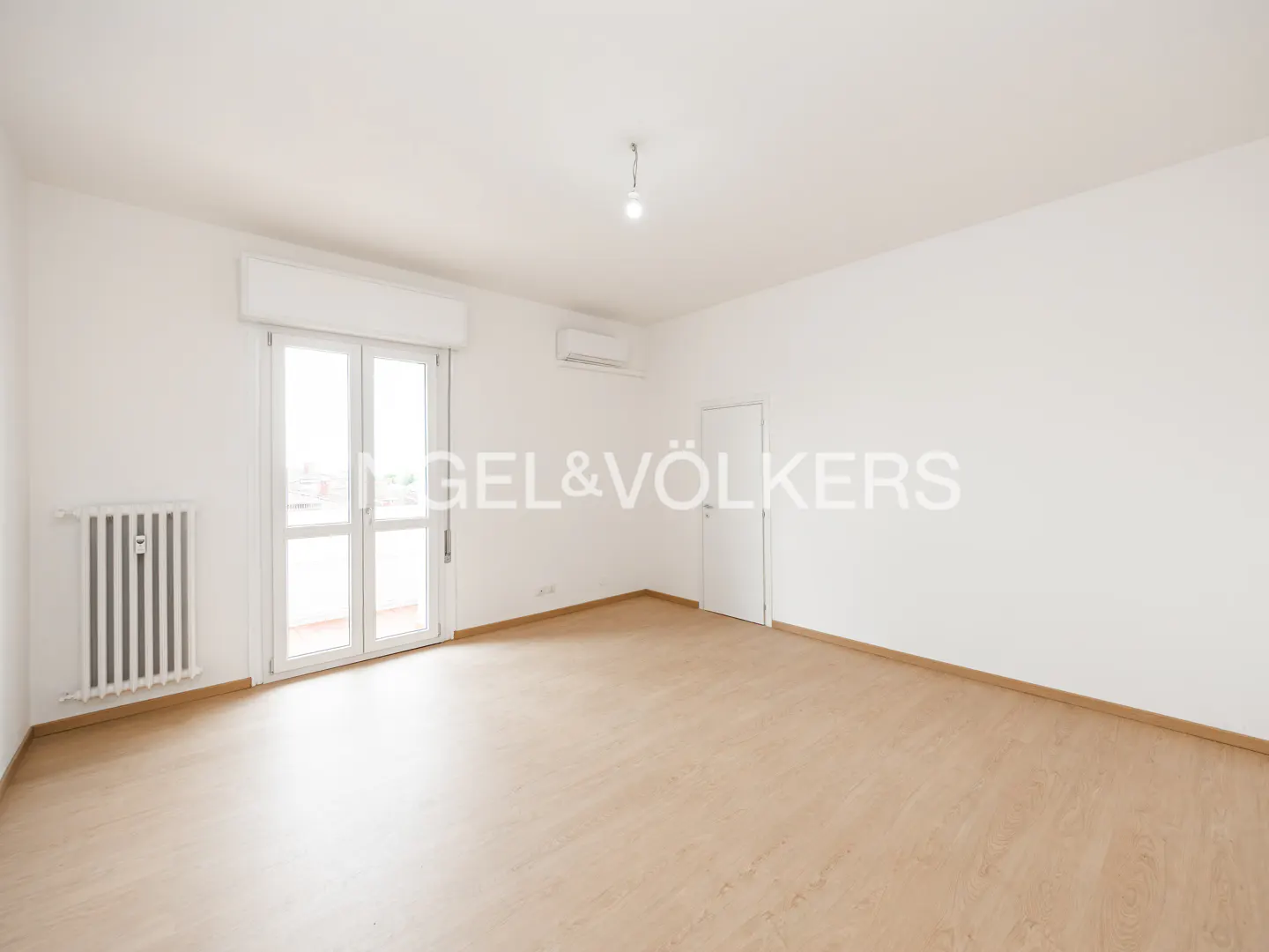 Bright, empty room with light wood floors, white walls, radiator, and balcony doors. Engel & Völkers logo visible.