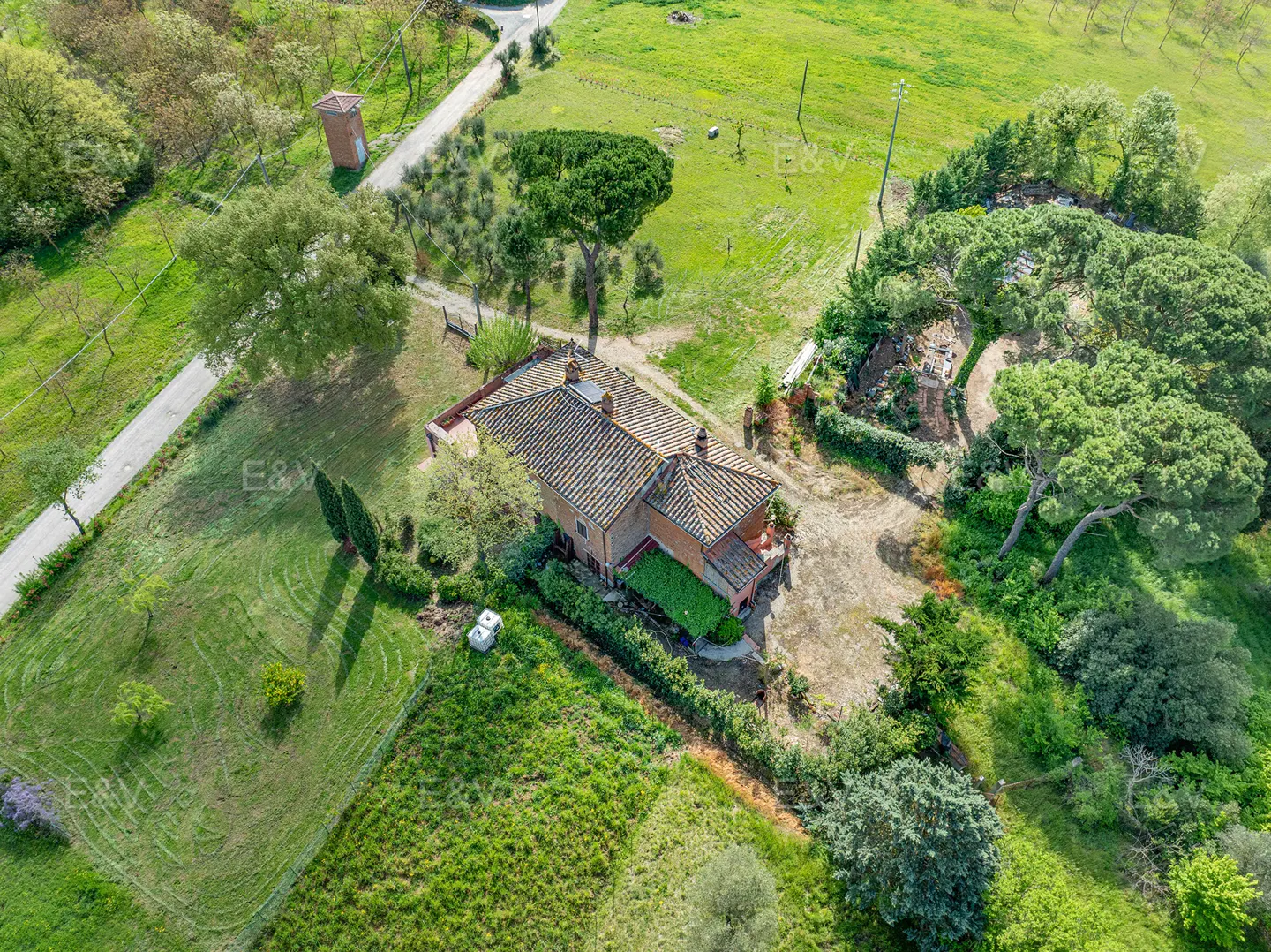Aerial view of a terracotta-roofed house surrounded by green fields and trees on a sunny day.